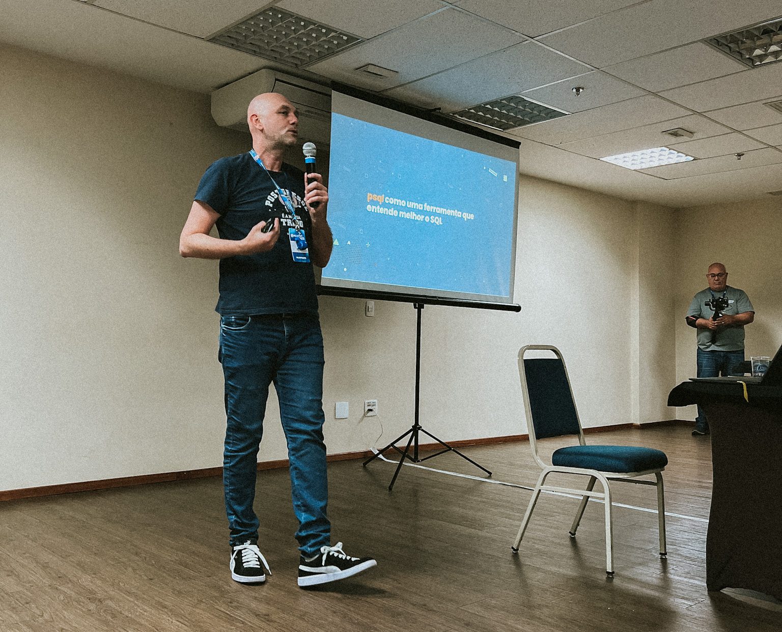 Dickson Guedes, consultor da Timbira, palestrando na PGConf.Brasil 2022. Ele segura um microfone enquanto apresenta sobre automação com SQL para uma plateia.